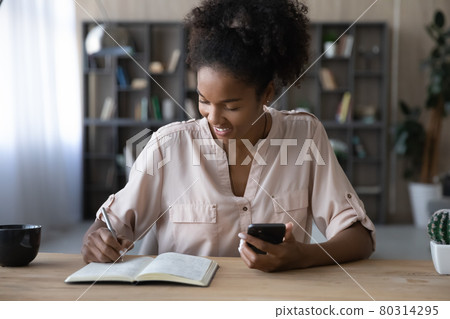 Smiling African American woman holding phone, taking notes in notebook Smiling African American woman holding phone, taking notes in notebook 80314295