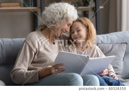 Old grandmother and preteen girl granddaughter read book on sofa Old grandmother and preteen girl granddaughter read book on sofa 80326350