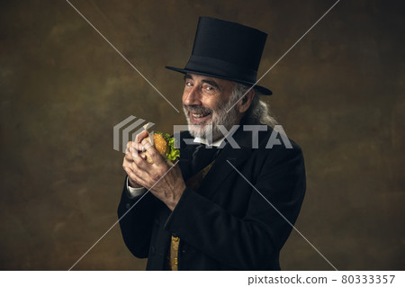 Handsome elderly gray-haired man, gentleman, aristocrat or actor eating fast food isolated on dark vintage background. Retro style, comparison of eras concept. 80333357