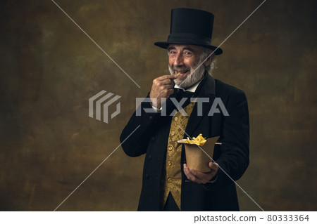 Handsome elderly gray-haired man, gentleman, aristocrat or actor eating fast food isolated on dark vintage background. Retro style, comparison of eras concept. 80333364