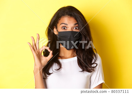 Concept of coronavirus, pandemic and lifestyle. Close-up of amazed and satisfied african-american girl, wearing black face mask, showing okay sign, like something good, yellow background Concept of coronavirus, pandemic and lifestyle. Close-up of amazed and satisfied african-american girl, wearing black face mask, showing okay sign, like something good, yellow background 80337111