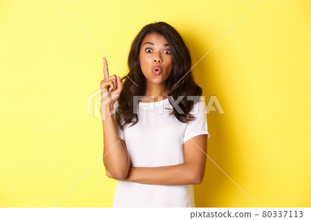 Portrait of smart african-american female student having a solution, suggesting idea, raising finger and looking excited, standing over yellow background Portrait of smart african-american female student having a solution, suggesting idea, raising finger and looking excited, standing over yellow background 80337113