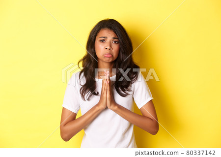 Portrait of sad african-american girl pleading, begging for help and sulking upset, standing over yellow background Portrait of sad african-american girl pleading, begging for help and sulking upset, standing over yellow background 80337142