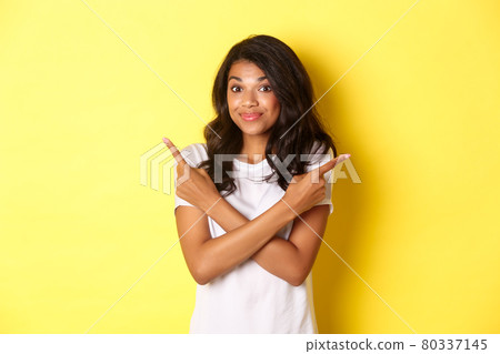 Portrait of cute indecisive african american girl, pointing fingers sideways and shrugging, asking advice with choices, standing over yellow background Portrait of cute indecisive african american girl, pointing fingers sideways and shrugging, asking advice with choices, standing over yellow background 80337145