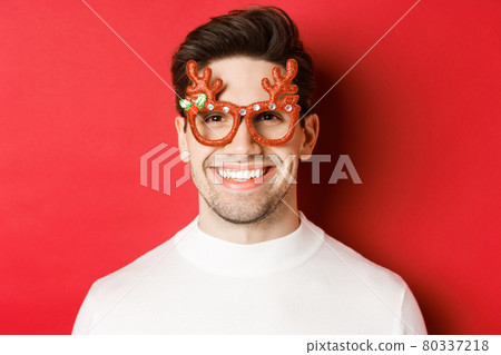 Concept of winter holidays, christmas and celebration. Close-up of attractive brunette guy in party glasses, smiling and looking happy, standing against red background 80337218