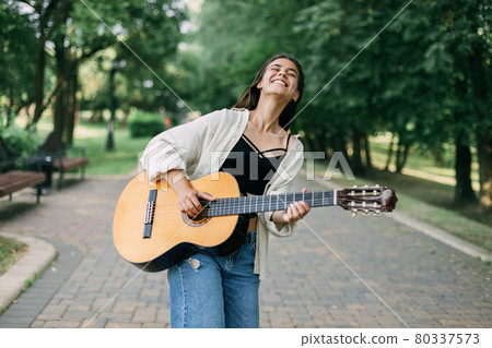 A cheerful musician girl enjoys playing the guitar in the park and laughs. Music, musician, guitar, freedom 80337573
