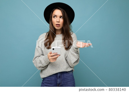Beautiful young brunette woman thinking wearing black hat and grey sweater holding smartphone 80338140