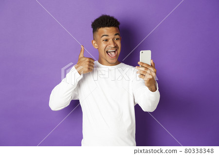 Image of smiling african-american male blogger, showing thumbs-up at mobile phone camera, recommend something, standing with smartphone over purple background 80338348