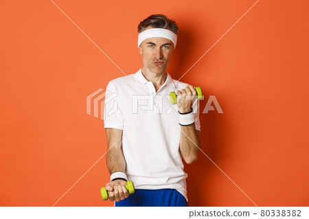 Portrait of cheeky middle-aged guy training in gym, lifting dummbells and smiling, workout against orange background 80338382