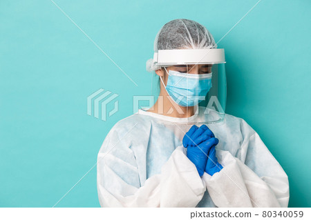 Concept of covid-19, pandemic and quarantine. Close-up of hopeful female doctor in personal protective equipment, holding hands in pray, begging or pleading, standing over blue background 80340059