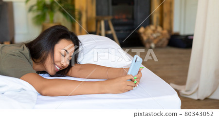 Happy laughing Asian woman lies in bed with mobile phone. She received a declaration of love from her boyfriend. Format photo 1x2. Happy laughing Asian woman lies in bed with mobile phone. She received a declaration of love from her boyfriend. Format photo 1x2. 80343052