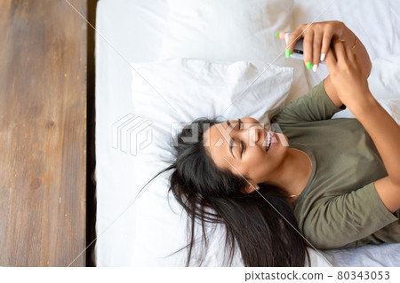 A happy laughing asian woman lies in bed with cell phone. A happy laughing asian woman lies in bed with cell phone. 80343053