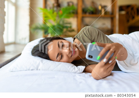 An asian woman lies in bed with cell phone. She woke up and was checking social media posts. 80343054