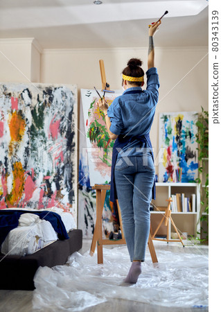 Full length of female artist raised her hand with paintbrush in it while thinking of a new painting, creating at home studio workshop Full length of female artist raised her hand with paintbrush in it while thinking of a new painting, creating at home studio workshop 80343139