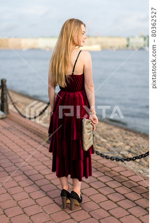 Slender blonde girl in elegant burgundy ankle-length dress turned her back on embankment. 80362527
