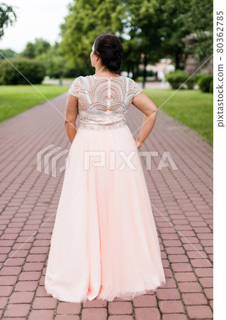 Brunette caucasian model in long pastel pink dress turned her back in park. 80362785