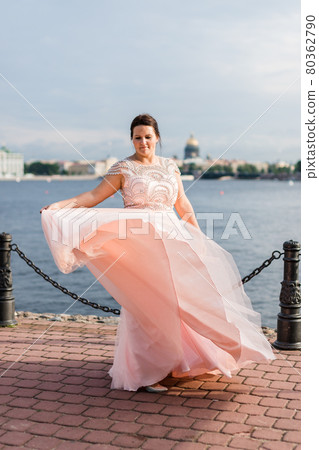 Pretty brunette caucasian model waving hem of pastel pink beaded dress on quay. 80362790