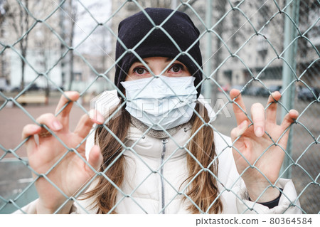 The girl in the medical mask is in the street in isolation and is holding fence. The girl in the medical mask is in the street in isolation and is holding fence. 80364584