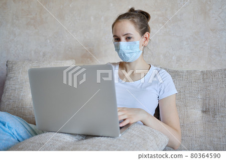 A girl in a medical mask is in quarantine at home working on a laptop. A girl in a medical mask is in quarantine at home working on a laptop. 80364590