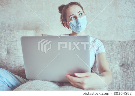 A girl in a medical mask is in quarantine at home holding a laptop and look out the window. 80364592