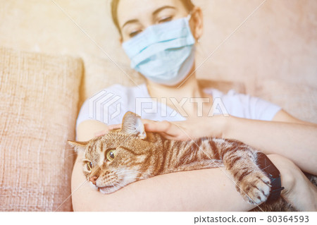 A girl in a medical mask is in quarantine at home petting a cat in her arms. 80364593