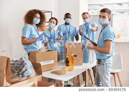 Group of diverse people wearing blue uniform, protective masks and gloves showing thumbs up while sorting donated food items, volunteering in community together 80369176