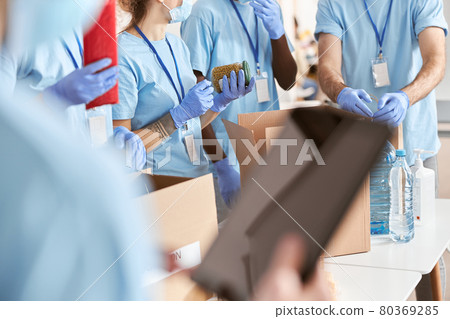 Cropped shot of volunteers in blue uniform, protective masks and gloves sorting, packing food and water in cardboard boxes, working together on donation project Cropped shot of volunteers in blue uniform, protective masks and gloves sorting, packing food and water in cardboard boxes, working together on donation project 80369285