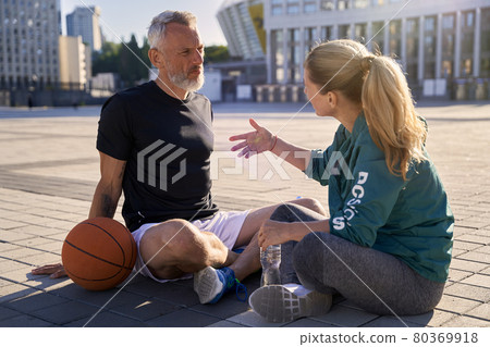 Middle aged couple, active man and woman in sportswear talking while relaxing after basketball workout outdoors on a sunny day Middle aged couple, active man and woman in sportswear talking while relaxing after basketball workout outdoors on a sunny day 80369918