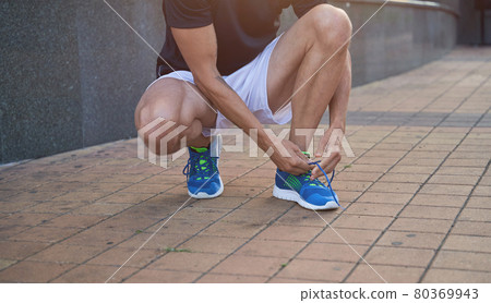 Close up shot of sportsman tying shoelaces on his blue running sneakers while getting ready for workout outdoors Close up shot of sportsman tying shoelaces on his blue running sneakers while getting ready for workout outdoors 80369943