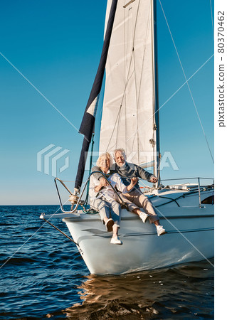 Full length of a beautiful and happy senior family couple embracing and smiling at camera while relaxing on a sail boat or yacht deck floating in a calm blue sea 80370462