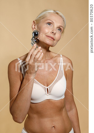 Beautiful mature blonde woman in underwear with glowing skin using metal face roller, looking away, posing isolated over beige background Beautiful mature blonde woman in underwear with glowing skin using metal face roller, looking away, posing isolated over beige background 80370620