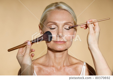Close up of hands of skillful artist doing natural makeup for mature blonde woman isolated over beige background. The woman closed her eyes with serenity 80370692