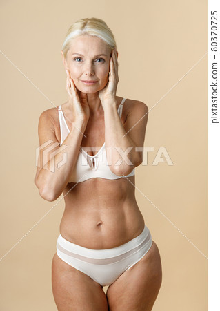 Woman in underwear with fit body looking at camera and touching facial skin, standing isolated over beige background 80370725