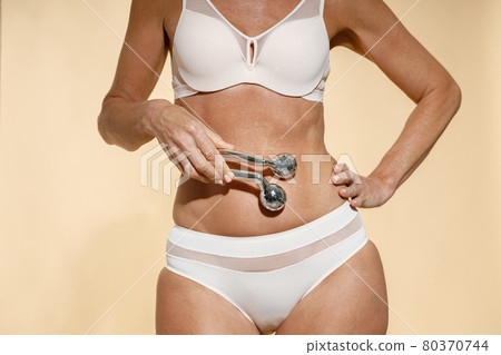 Cropped shot of fit mature blonde woman in underwear holding metal face roller over her stomach, posing isolated over beige background 80370744