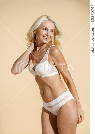 Confident caucasian mature blonde female model with fit body in white lingerie smiling aside, posing half naked isolated over beige background 80370747
