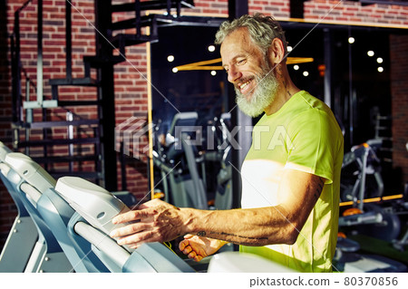 Side view of a happy middle aged athletic man in sportswear adjusting speed on a treadmill while having cardio workout in a gym or sport club 80370856