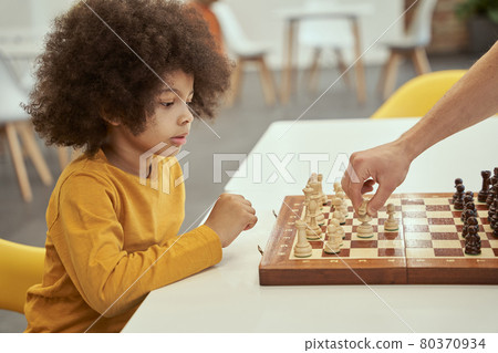 Enthusiastic player. Cute little clever boy thinking while playing chess with adult, sitting at the table indoors Enthusiastic player. Cute little clever boy thinking while playing chess with adult, sitting at the table indoors 80370934