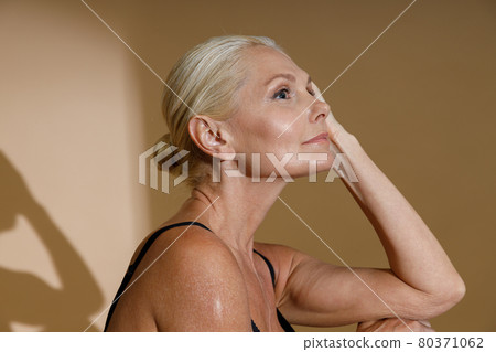Studio portrait of beautiful mature blonde woman in black underwear looking away while posing half naked isolated over beige background Studio portrait of beautiful mature blonde woman in black underwear looking away while posing half naked isolated over beige background 80371062