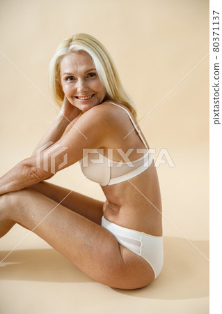 Studio shot of lovely mature blonde woman posing in underwear, sitting on the floor and smiling at camera isolated over beige background Studio shot of lovely mature blonde woman posing in underwear, sitting on the floor and smiling at camera isolated over beige background 80371137