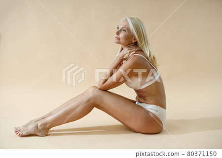 Side view of sensual blonde woman posing in underwear, sitting on studio floor and looking away isolated over beige background 80371150