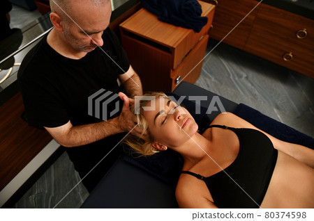 Beautiful woman lying on massage table with eyes closed and receiving professional medical therapeutic head massage given by masseur at health spa 80374598