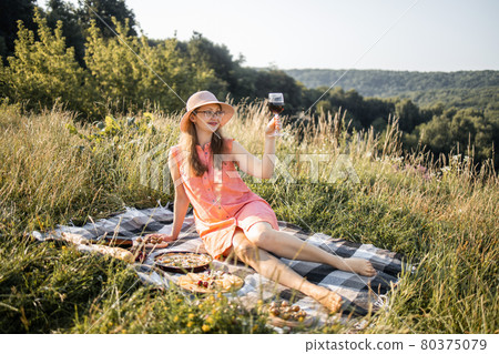 Charming lady with glass of red wine sitting on the blanket outdoors 80375079