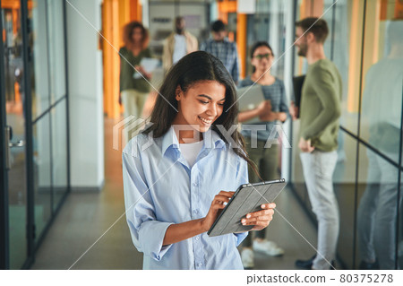 Mirthful young woman using tablet while her colleagues chatting Mirthful young woman using tablet while her colleagues chatting 80375278