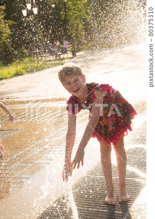 Happy girl in the spray of the fountain. The child smiles in the rays of the sun. Summer heat 80375310