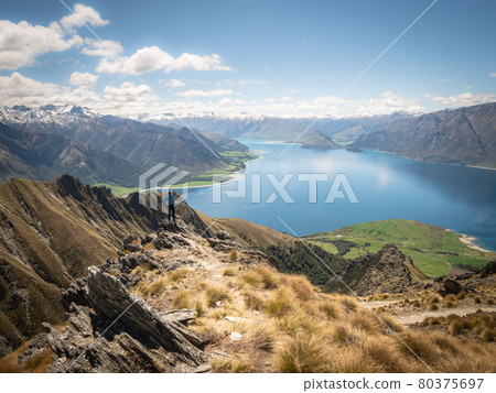Man standing on the edge of rock enjoying beautiful vista with lush green meadows, blue lake and snowy mountains, sunny day on Isthmus Peak summit Man standing on the edge of rock enjoying beautiful vista with lush green meadows, blue lake and snowy mountains, sunny day on Isthmus Peak summit 80375697