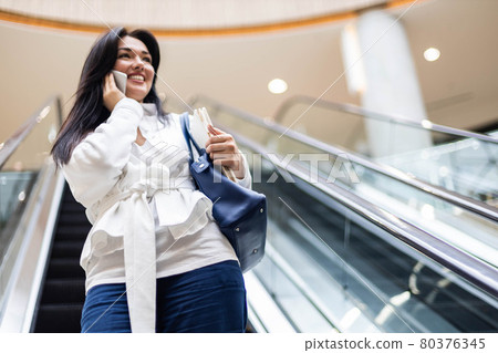 Cheerful business woman talking smartphone smiling having positive emotion at modern shopping mall Cheerful business woman talking smartphone smiling having positive emotion at modern shopping mall 80376345