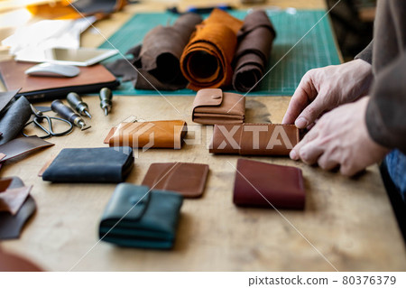 Craftsman hands laying out leatherwork on wooden table at leather workshop. Handwork accessories 80376379