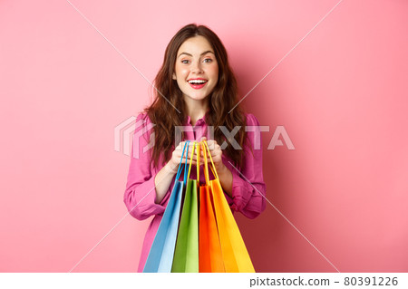 Young beautiful lady going on shopping, holding bags and smiling excited, buying with discounts, standing against pink background Young beautiful lady going on shopping, holding bags and smiling excited, buying with discounts, standing against pink background 80391226