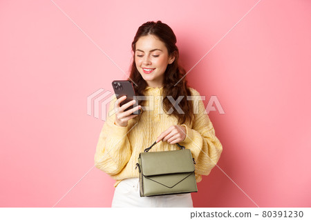 Glamour girl holding purse and texting message on smartphone, reading mobile phone screen with carefree smile, standing over pink background Glamour girl holding purse and texting message on smartphone, reading mobile phone screen with carefree smile, standing over pink background 80391230