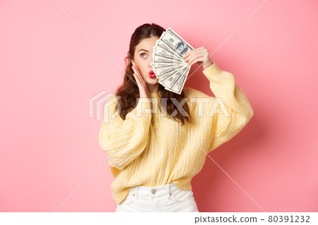Attractive female model showing money, holding dollar bills on half of face, touching cheek and making kissing lips, standing against pink background Attractive female model showing money, holding dollar bills on half of face, touching cheek and making kissing lips, standing against pink background 80391232